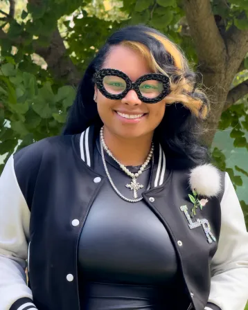 Damaris standing in front of a tree near Clothier Hall wearing a black shirt and black and white jacket with black glasses.