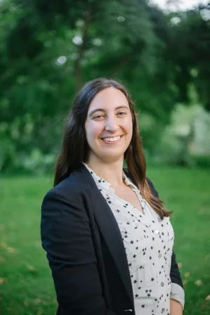 Image of Kristen Recine. She is a white woman with mid-length brown hair. She is smiling at the camera with a green background of trees. She is wearing a black jacket and a white shirt with stars on it.