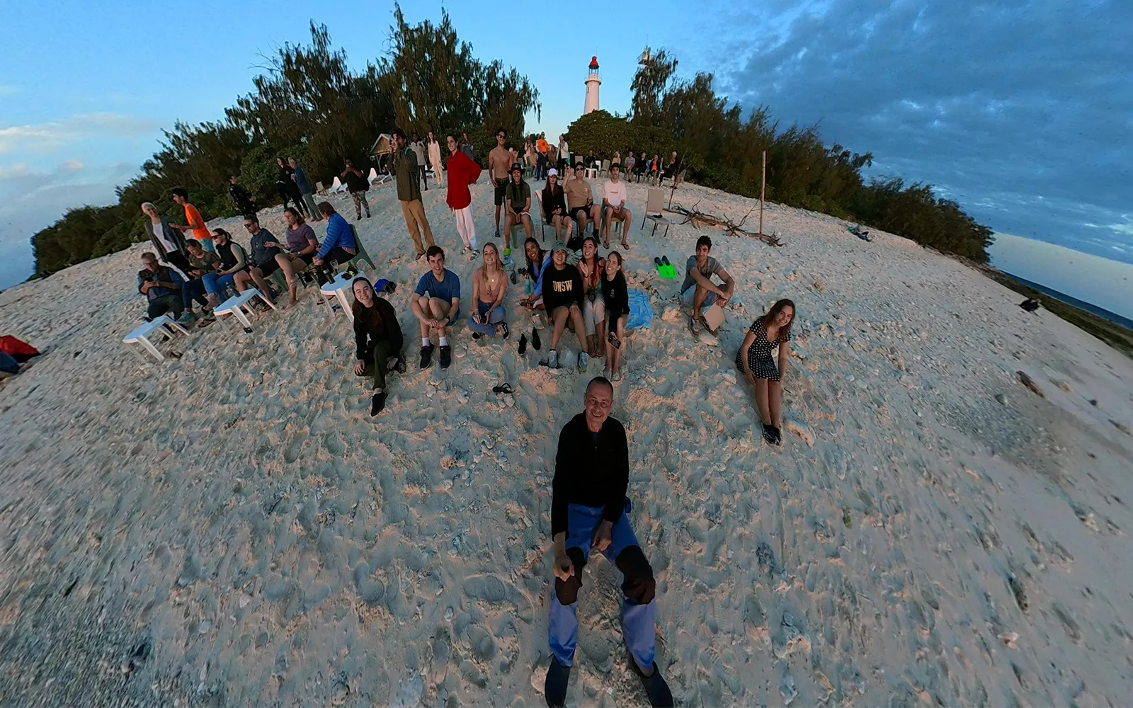 Group of students sitting on beach (facing camera) with lighthouse in the background