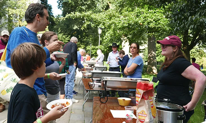 A man and a child stand near a table and talk to the woman standing on the other side of the table