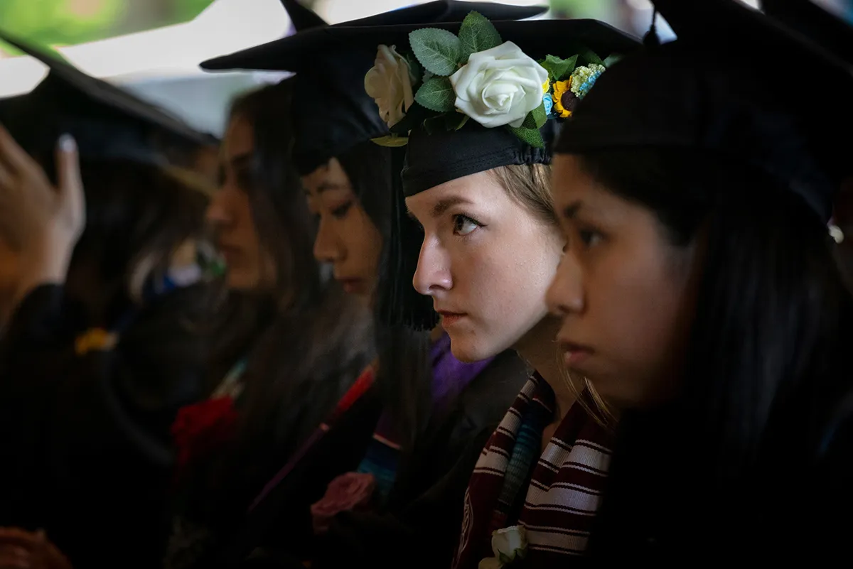 seated student wearing cap and gown looks toward stage