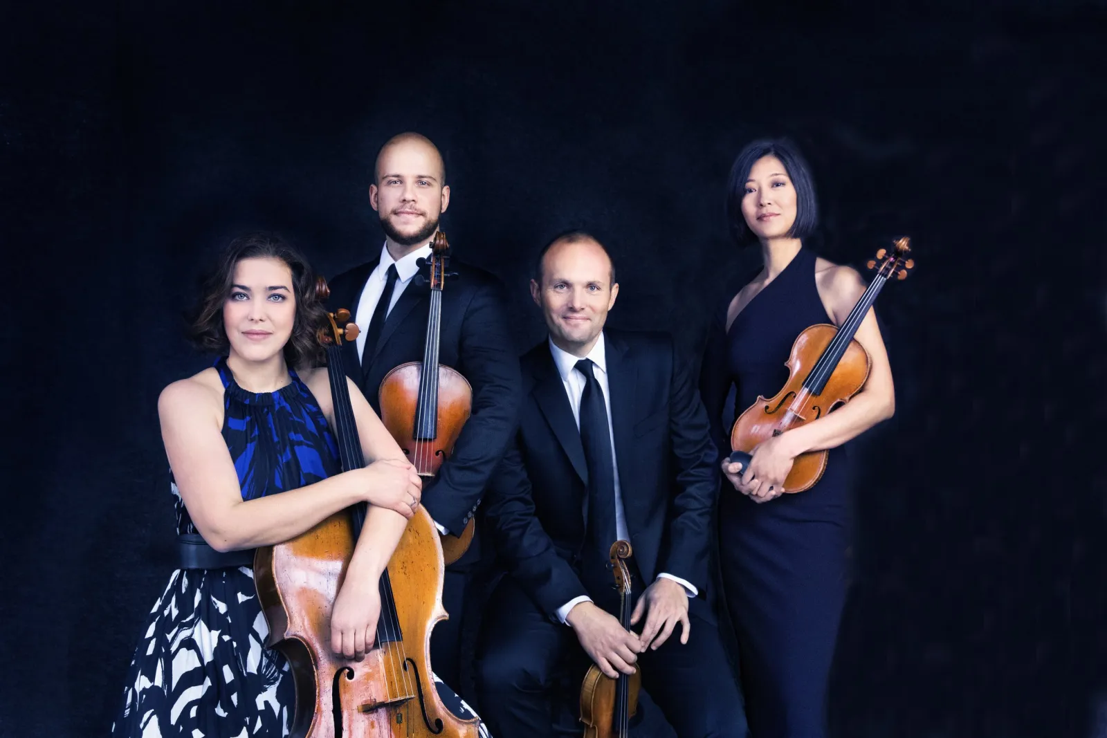 Four musicians posing with their string instruments in front of a black background