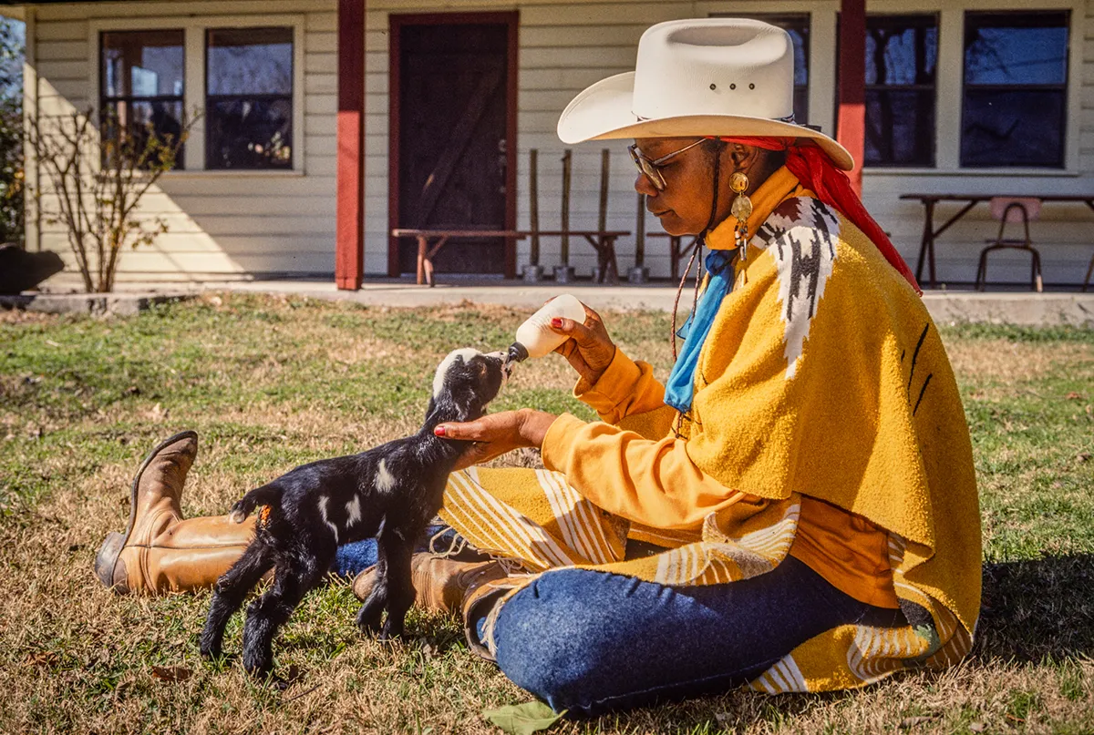 Woman in cowboy hat feeds kid goat with bottle