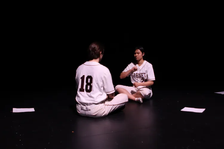 Photograph from Night of Scenes 2023. Two individuals sit across from each other with their legs crossed. They are wearing baseball uniforms. We see the back of one of the figures whose jersey reads the number eighteen. The other figure's jersey reads "GARNET" and they are holding their fist to their chest. On the floor around the two are sheets of paper 