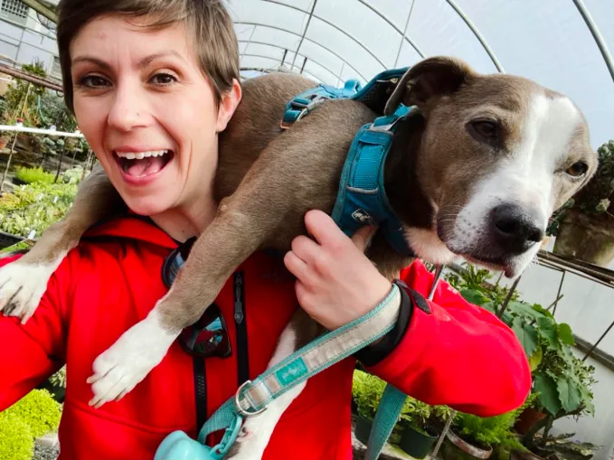 Missy Furth stands in a greenhouse and holds a dog over her shoulder.