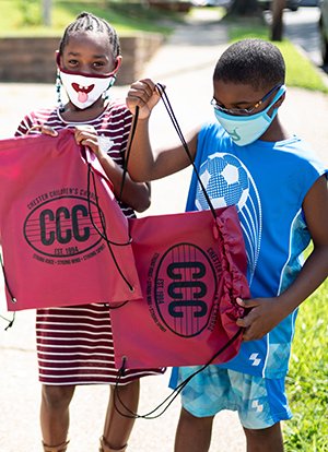 Two children wearing masks hold red drawstring bags on sidewalk.