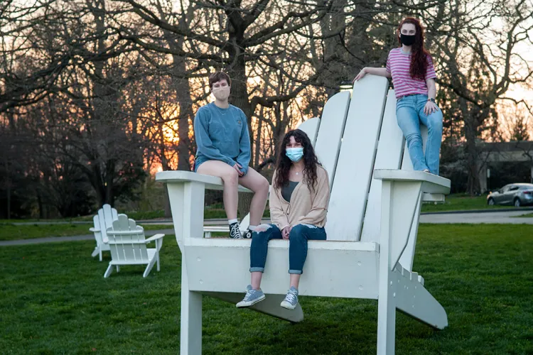 Three students wearing masks sit on oversized white Adirondack chair during sunset