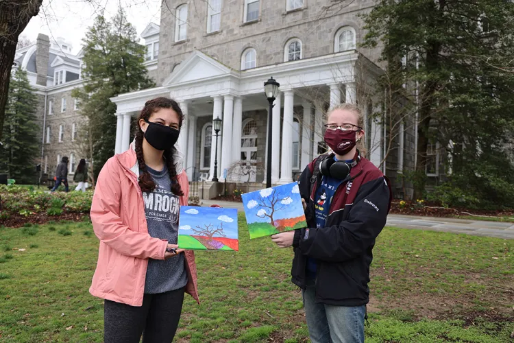 Two students wearing masks hold up paintings of bare trees.