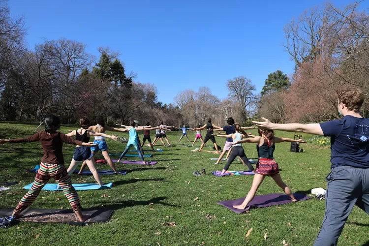 Students practice yoga outdoors, posing with legs wide and arms outstretched. 