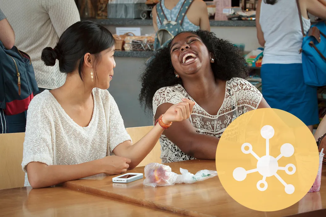 Two people sitting and laughing at table. Yellow circle featuring symbol of network in bottom right corner.