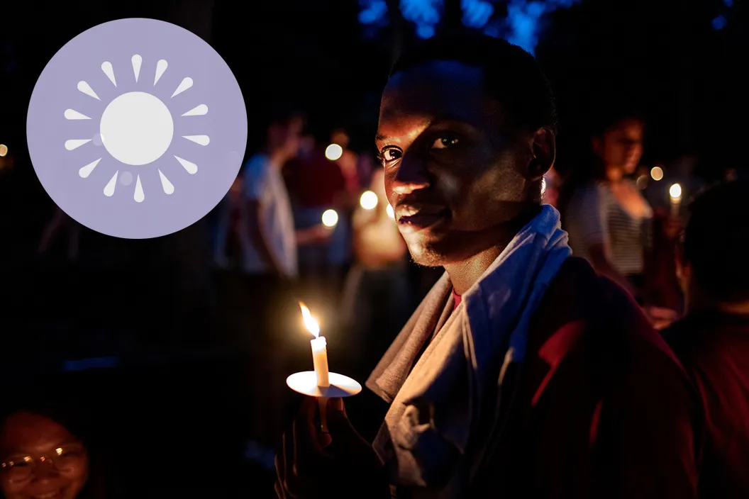 Person holds candle outdoors during evening. Purple logo featuring symbol of sun in upper left corner.