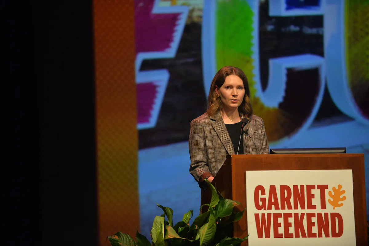 A woman stands behind a podium in front of a large screen. She has red hair and is wearing a brown checked blazer. There is a tropical plant next to the podium. A sign reading "Garnet Weekend" in red text is mounted on the podium.